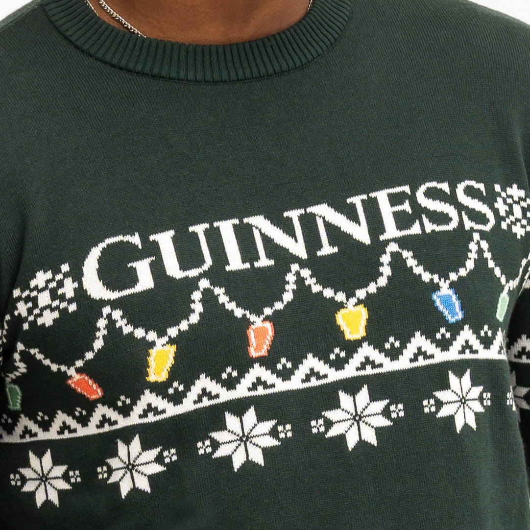 Someone wearing the Guinness Holiday Pint Lights Sweater - Green from Guinness Webstore US, featuring "GUINNESS," festive pint-shaped lights, and white snowflakes.