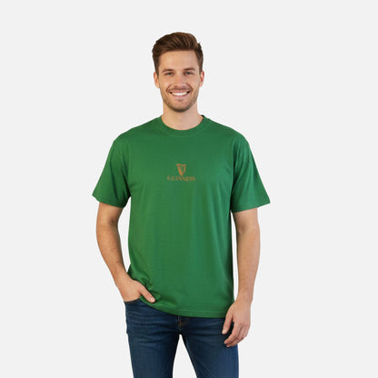 A man stands smiling with one hand in his pocket, wearing the Guinness Are you Toucan to Me Tee in green from Guinness Webstore US and blue jeans, set against a plain white background.