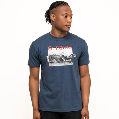 A person stands against a plain background wearing a Guinness Transport Collection Navy Tee by Guinness and black pants, looking slightly to the side.