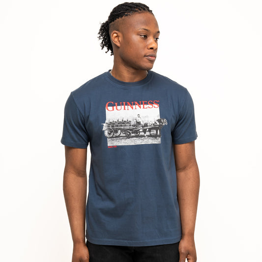 A person stands against a plain background wearing a Guinness Transport Collection Navy Tee by Guinness and black pants, looking slightly to the side.