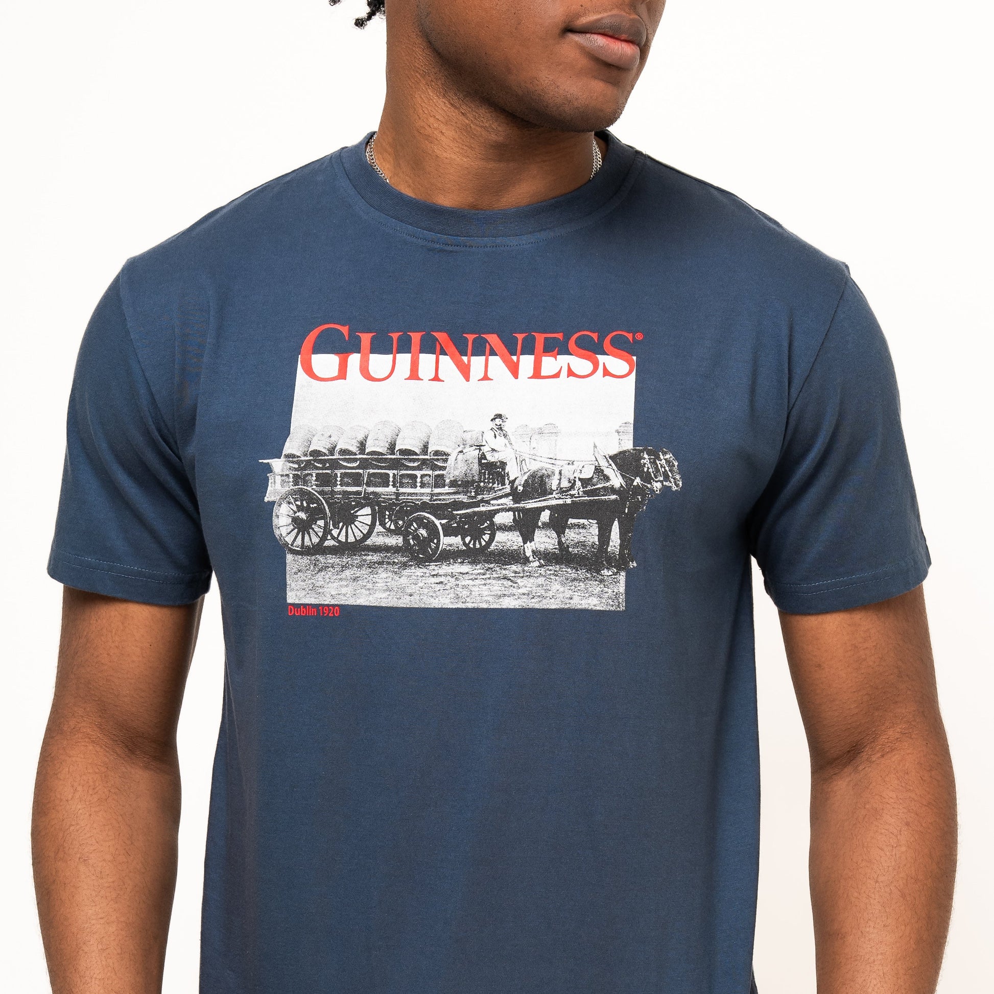 A person wearing the Guinness Transport Collection Navy Tee, a 100% cotton shirt featuring a vintage horse-drawn cart graphic and the iconic red Guinness Trademark Harp Logo.