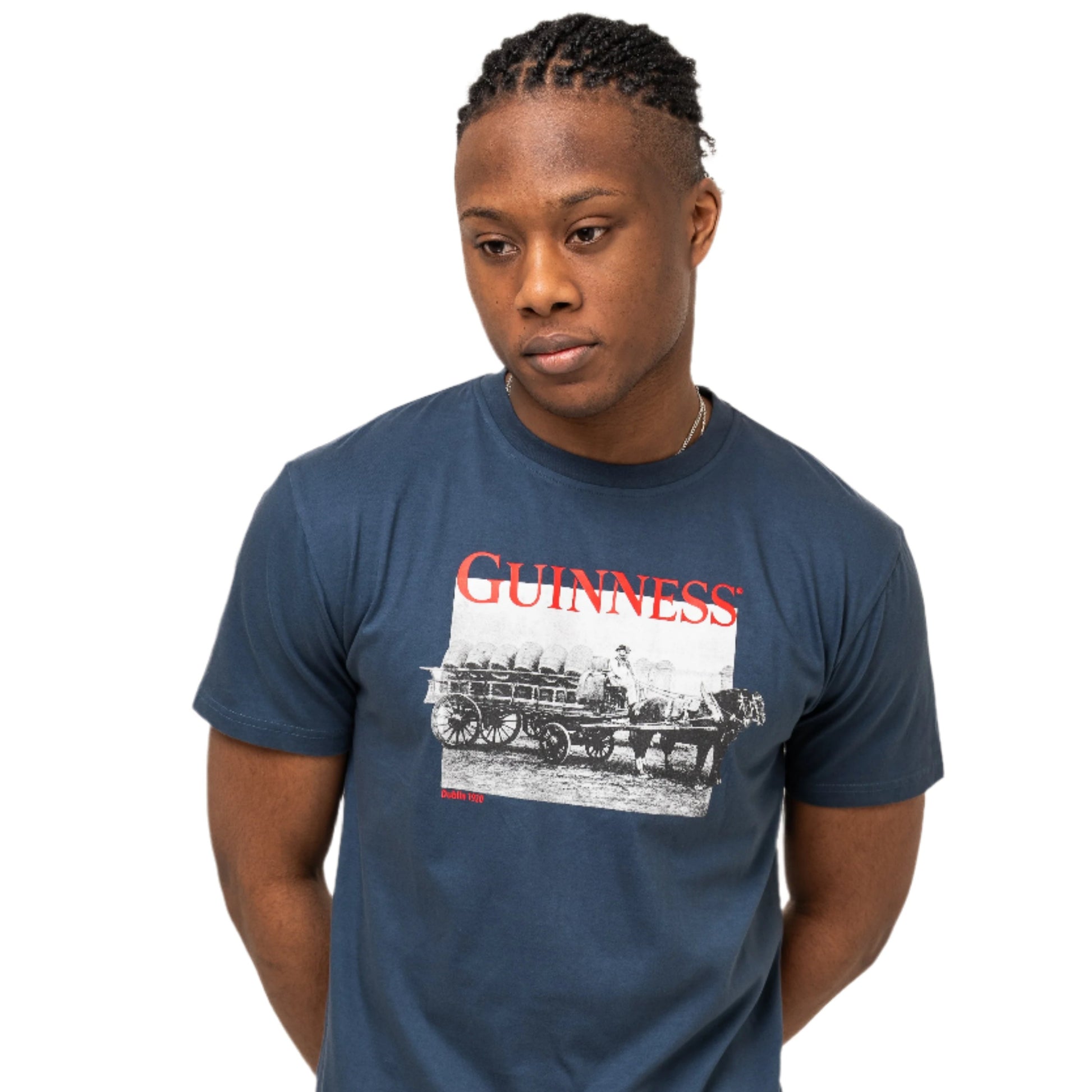 A person stands against a plain background wearing a Guinness Transport Collection Navy Tee by Guinness, made from 100% cotton and featuring a vintage train graphic with the iconic Harp Logo, paired with black pants.
