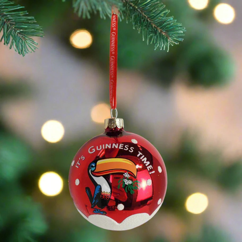 A Guinness Toucan Glass Bauble, boasting a vibrant red color with an endearing toucan illustration and the phrase "It's Guinness Time," hangs festively from a tree branch.