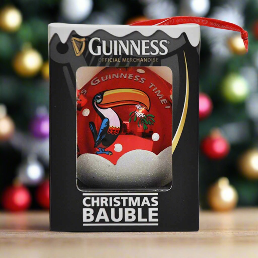 The Guinness Toucan Glass Bauble features a toucan holding a pint, elegantly encased in a festive ornament. Softly blurred Christmas lights twinkle in the background, enhancing the holiday charm.