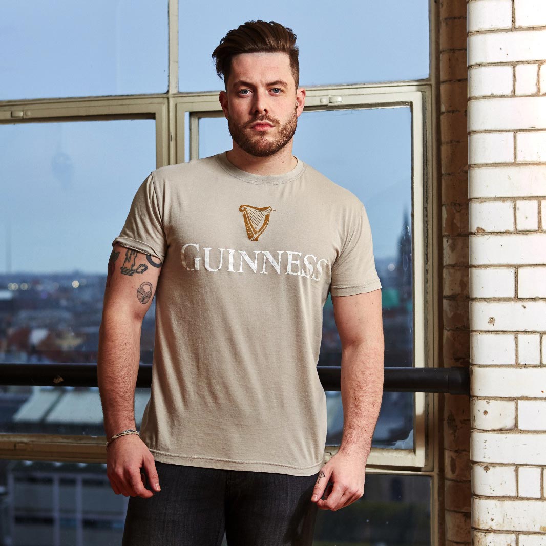 A cotton t-shirt enthusiast proudly stands in front of a window, showcasing his love for Guinness with the Guinness Trademark Label T-Shirt Beige prominently displayed on his shirt.