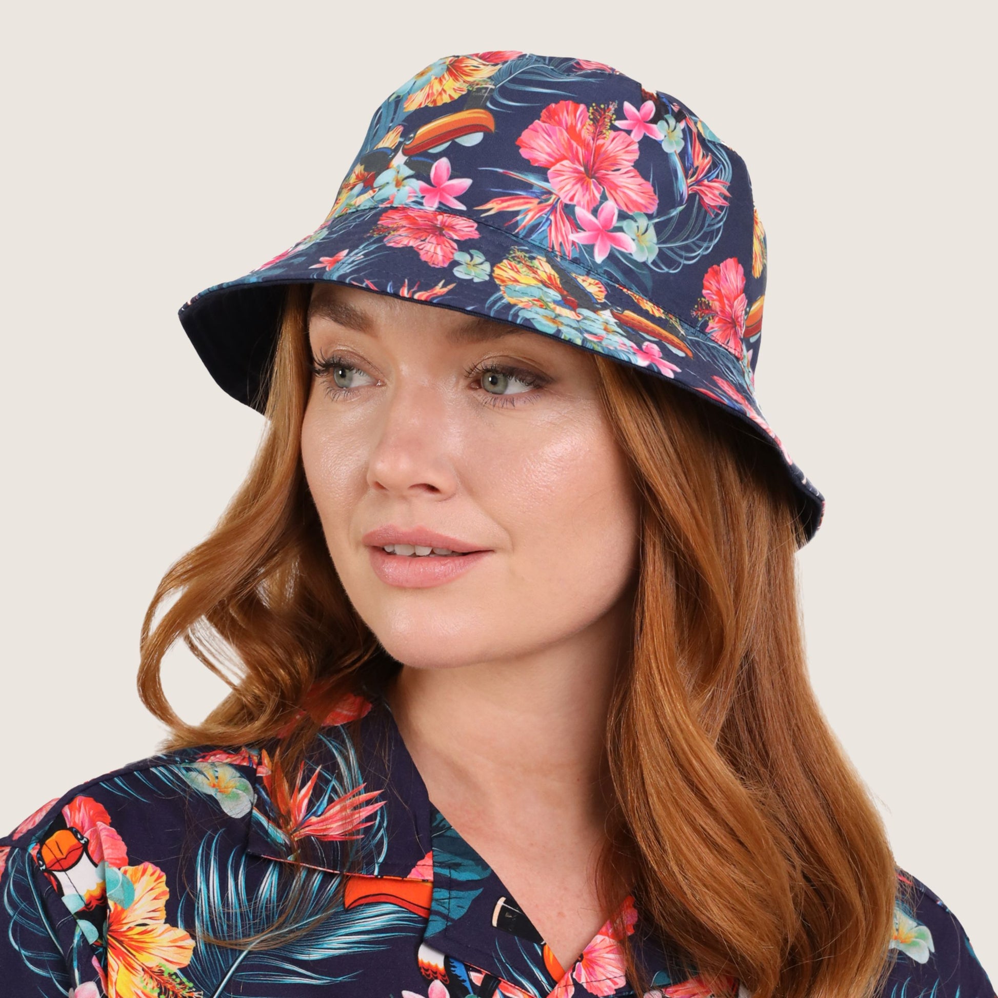 A woman with long red hair wears the Guinness Hawaiian Reversible Bucket Hat from Guinness Webstore US and a matching floral-patterned shirt, looking slightly to the side.