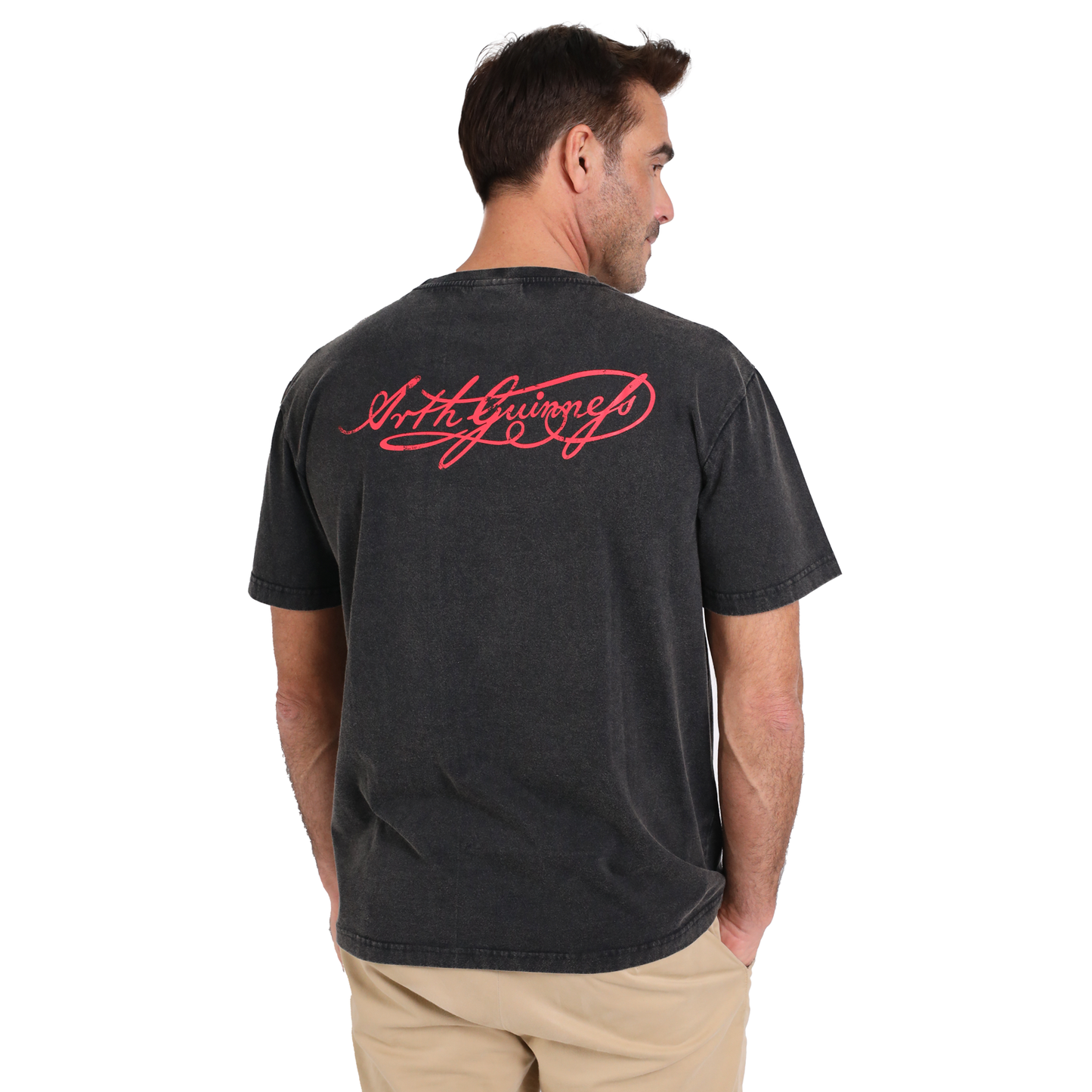 Man in a Guinness Black Distressed Label Tee with red cursive text on the back, facing sideways and looking to his left, wearing beige pants. The shirt features an Arthur Guinness signature print for a classic touch.