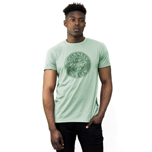 A man wearing a "Guinness Green Bottle Cap Tee" made by Guinness, made of cotton blend fabric.