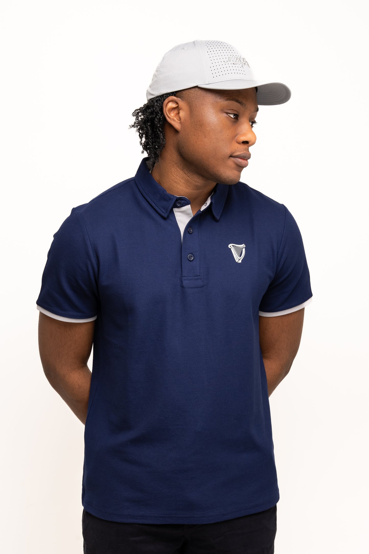 A person wearing a blue polo shirt with a chest emblem and the Guinness Performance Signature Cap from Guinness Webstore US stands against a plain white background with hands behind their back.