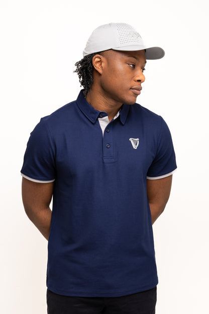 A person wearing a blue polo shirt with a chest emblem and the Guinness Performance Signature Cap from Guinness Webstore US stands against a plain white background with hands behind their back.