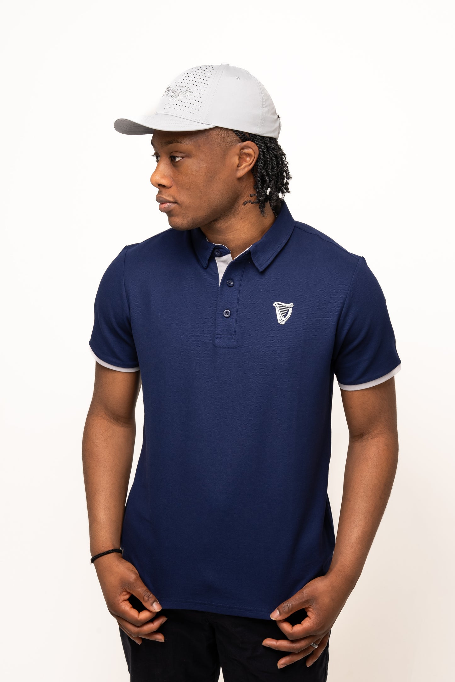 Wearing a navy blue polo with an embroidered logo and the Guinness Performance Signature Cap from Guinness Webstore US, a person stands against a plain white background, gazing to the side.