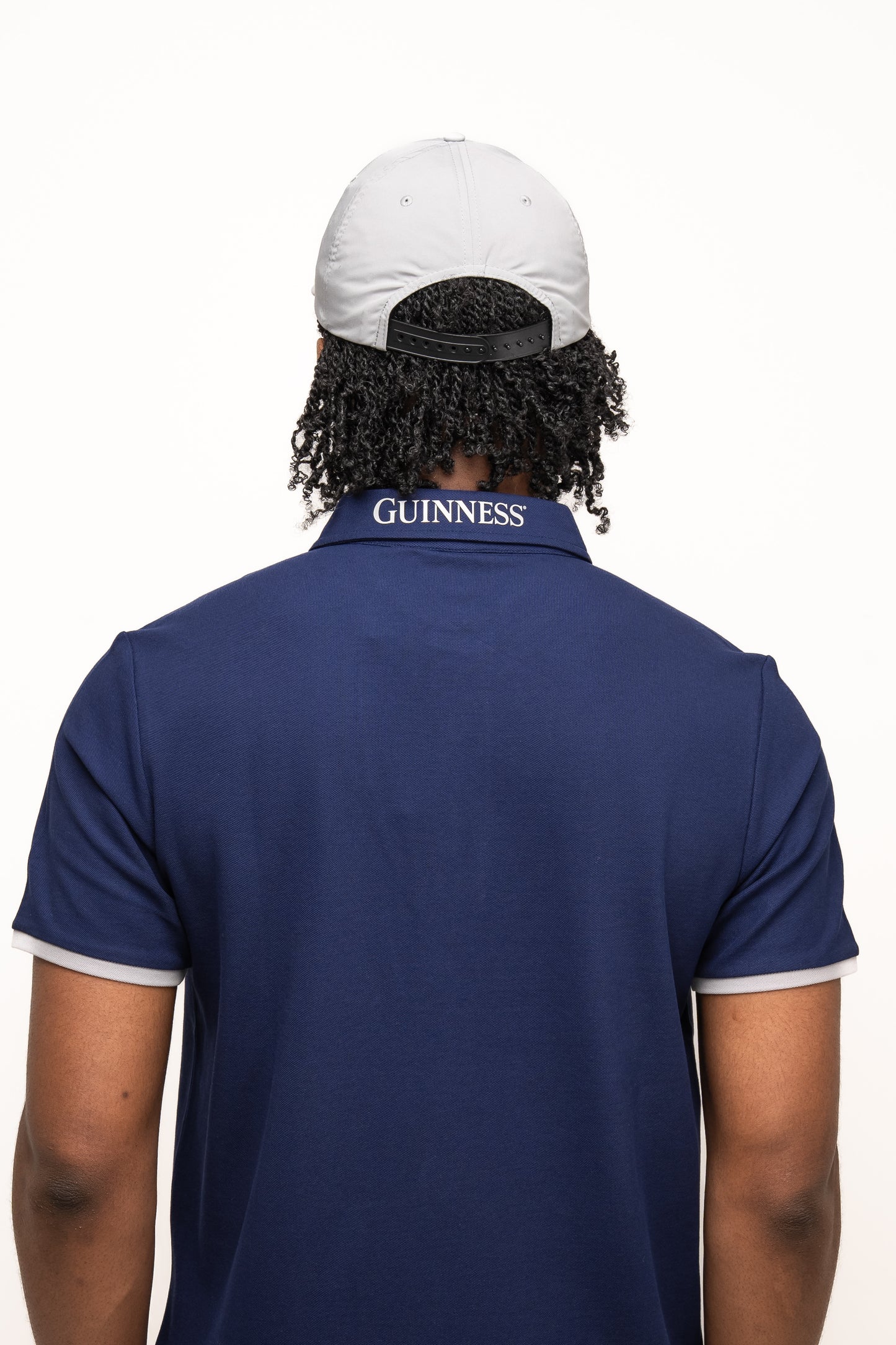 A person in a Guinness Performance Signature Cap from Guinness Webstore US and a navy blue polo with "GUINNESS" on the back collar, seen from behind against a white background.
