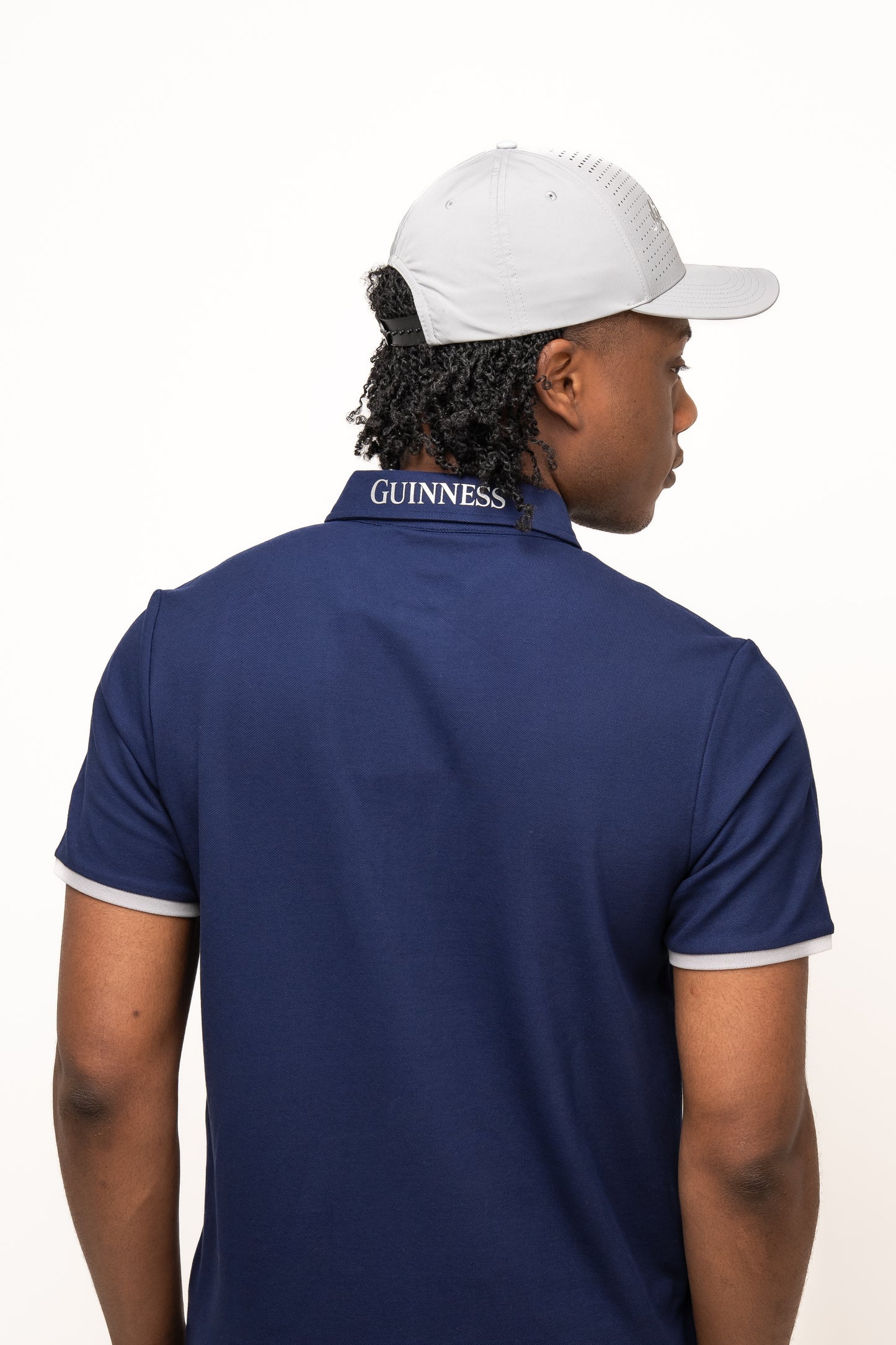 A person seen from behind wears a light gray Guinness Performance Signature Cap from Guinness Webstore US with quick-dry tech and a navy blue polo with "GUINNESS" on the collar, set against a plain white background.