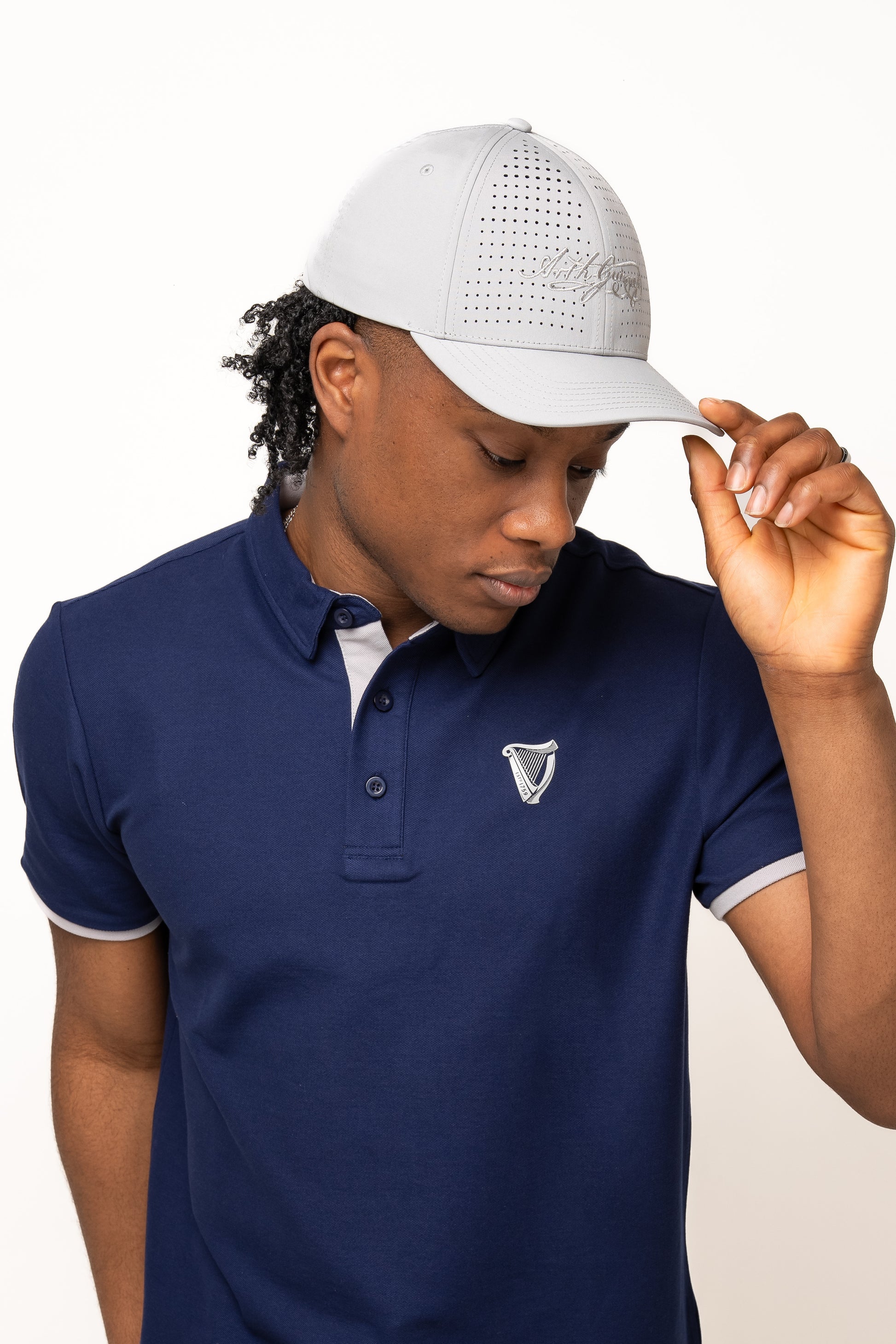 A person in a navy blue polo with a crest and a light gray Guinness Performance Signature Cap by Guinness Webstore US, featuring quick-dry technology, looks down while adjusting the cap with one hand.