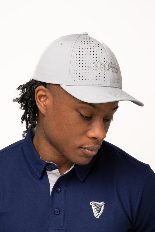 A person wearing a light gray Guinness Performance Signature Cap from Guinness Webstore US, featuring quick-dry technology, and a navy collared shirt with a white harp logo looks down against a plain background.