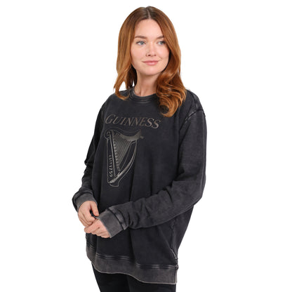 A woman with long auburn hair models the Guinness Embossed Print Trademark Sweater from Guinness Webstore US—an oversized black BCI cotton fleece sweatshirt with the embossed logo and harp, shown against a white background.