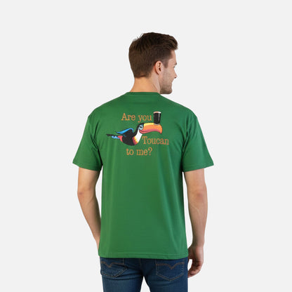 A man wears the Guinness Are you Toucan to Me Tee in green from Guinness Webstore US, made of BCI cotton and featuring a toucan graphic with a beer glass and "Are you Toucan to me?" on the back.