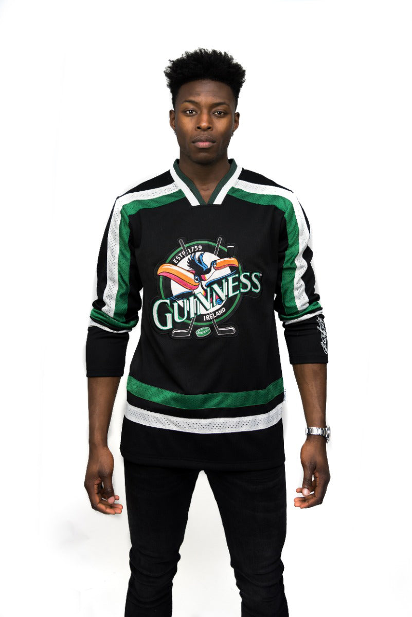 A man faces forward wearing the Guinness Toucan Hockey Jersey in black and green, featuring an embroidered toucan logo, with a plain white background.