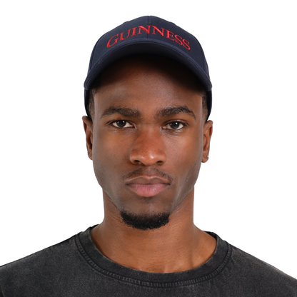 A man wearing a dark-colored Guinness Baseball Cap from the Transport Collection by Guinness Webstore US and a dark t-shirt looks directly at the camera with a neutral expression, embodying a timeless style.