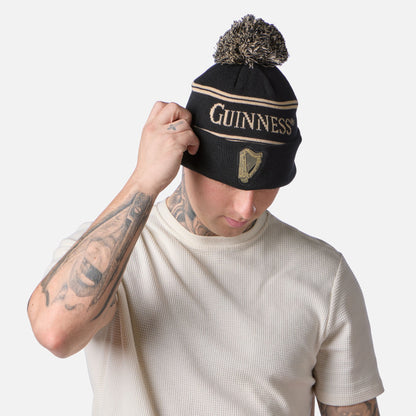 Wearing a cream textured shirt, a person adjusts their Guinness Black & Gold Sports Beanie – Vintage Harp Knit, looking down.