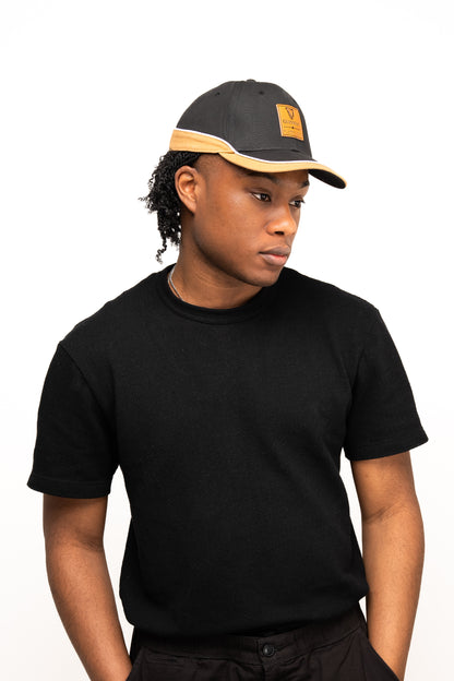 A person wearing a black t-shirt and the Guinness Black Cap with Caramel Edge from Guinness Webstore US stands against a plain white background, looking slightly to the side.