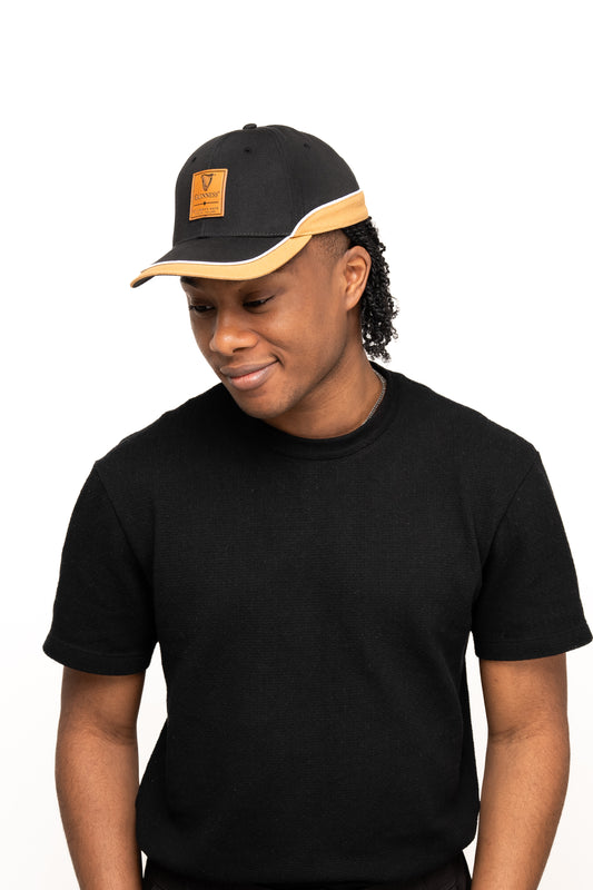 A person wearing a black t-shirt and the Guinness Black Cap with Caramel Edge from Guinness Webstore US stands against a plain white background, looking slightly downward.
