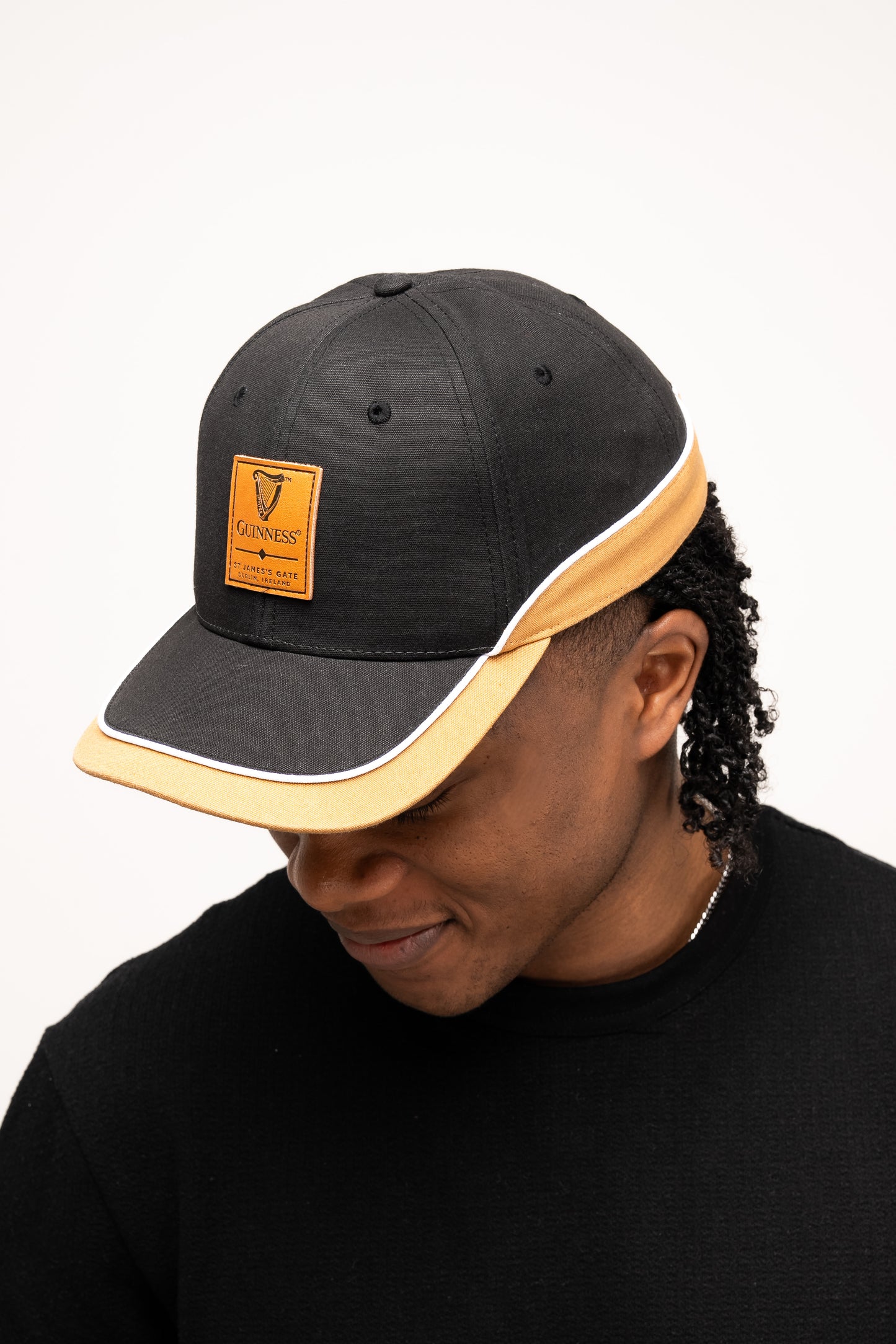 A person wears a Guinness Black Cap with Caramel Edge from Guinness Webstore US and a black sweater, looking downward with their face partially visible.