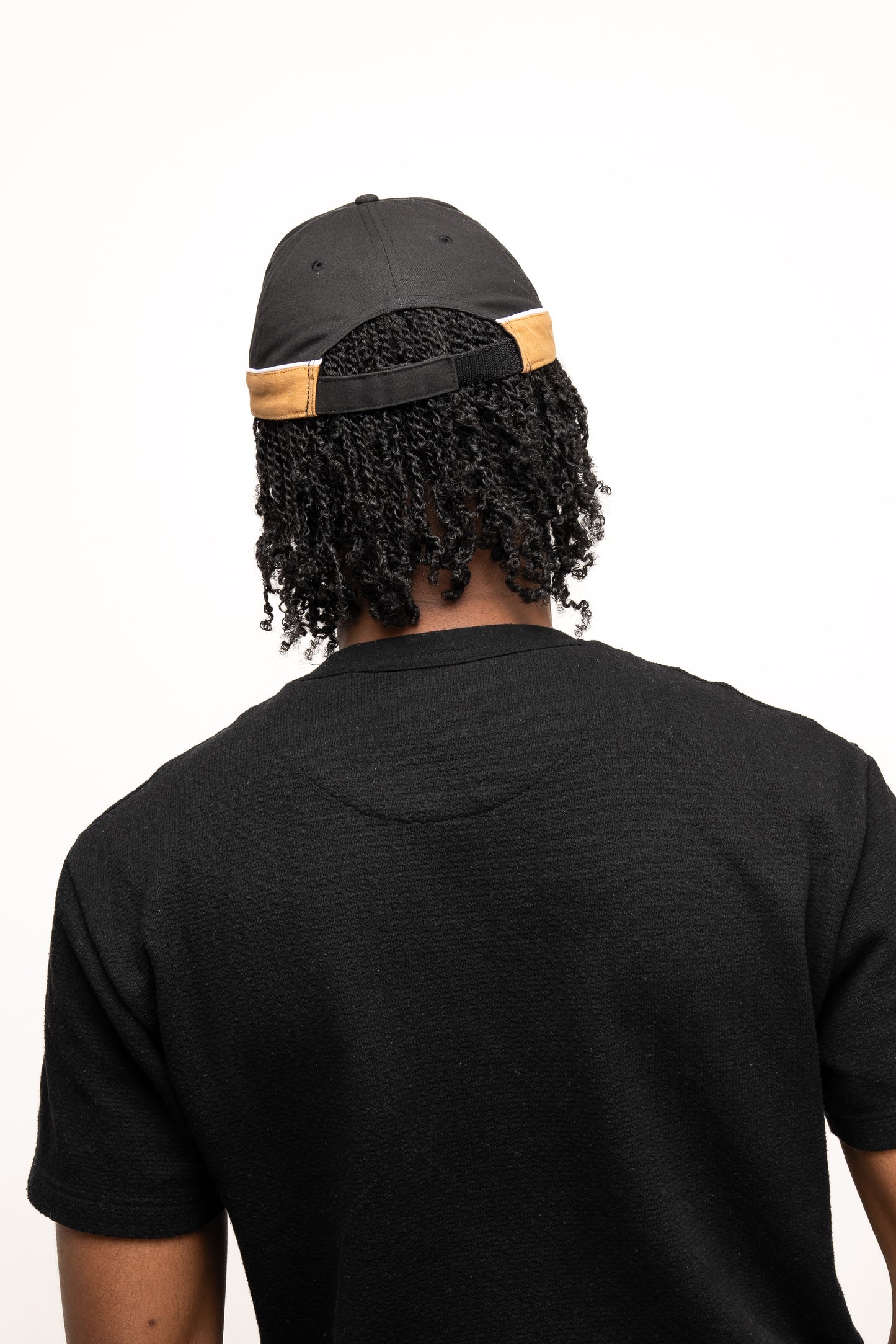 A person with curly hair wears the Guinness Black Cap with Caramel Edge from Guinness Webstore US and a black shirt, shown from the back against a plain white background.