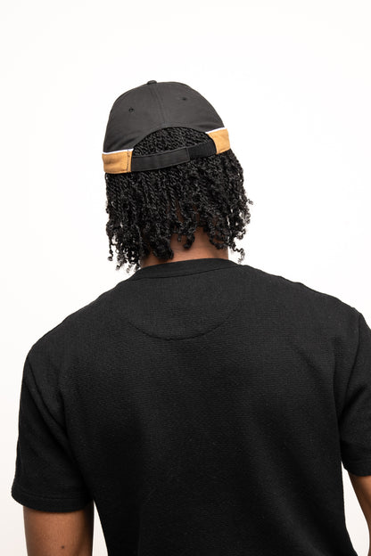 A person with curly hair wears the Guinness Black Cap with Caramel Edge from Guinness Webstore US and a black shirt, shown from the back against a plain white background.
