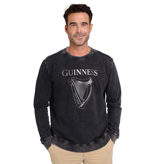 A man stands facing forward with his hands in his pockets, showcasing the Guinness Metallic Print Trademark Sweater from Guinness Webstore US, featuring a metallic harp logo and crafted from sustainable cotton.
