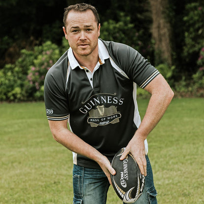 A man holding a rugby ball in a grassy field, wearing a moisture-wicking Made of More Rugby Jersey with Guinness branding.