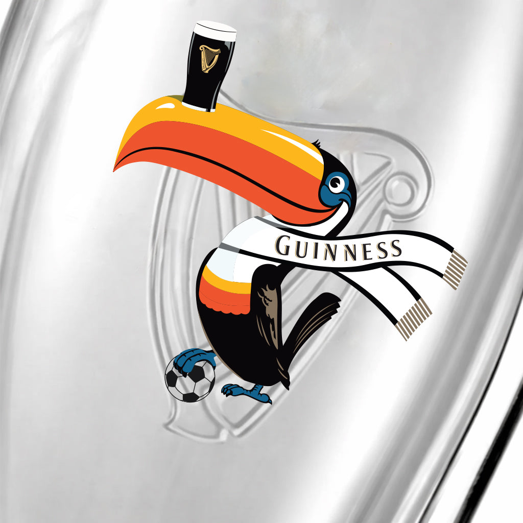 A toucan in a Guinness scarf balances a soccer ball on one foot and the Guinness Soccer Pint Glass – Special Edition on its beak, set against a silver background with a faint harp logo—celebrating the 2026 tournament for Guinness Webstore US.