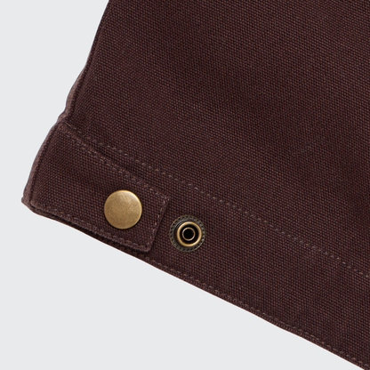 Close-up of a Guinness Cooperage Work Jacket corner in brown heavyweight cotton canvas with brass snap fasteners on a light background.
