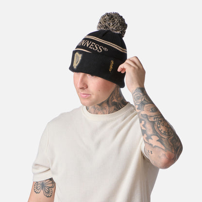 Man in a white shirt adjusts a Guinness Black & Gold Sports Beanie – Vintage Harp Knit; tattoos visible on his arms and neck.