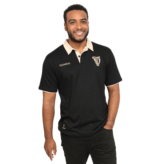 A man wearing a Guinness Black and Cream Traditional Short Sleeve Rugby, smiling, stands with one hand in his pocket against a plain white background.