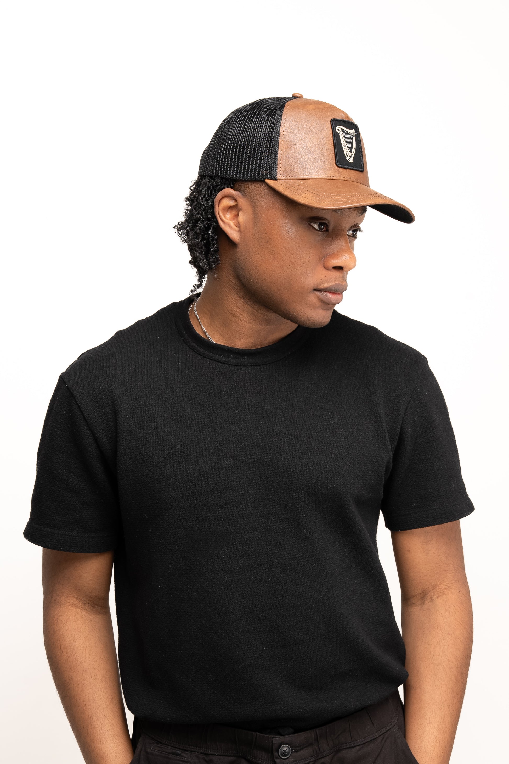 A person wears a black short-sleeve shirt and the Guinness Brown Leather Trucker Cap from Guinness Webstore US, featuring a leather peak and snap-back closure, while looking sideways against a plain white background.