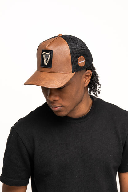A person wearing a black t-shirt and the Guinness Webstore US Guinness Brown Leather Trucker Cap with leather peak, harp logo, and badge, looking downward.