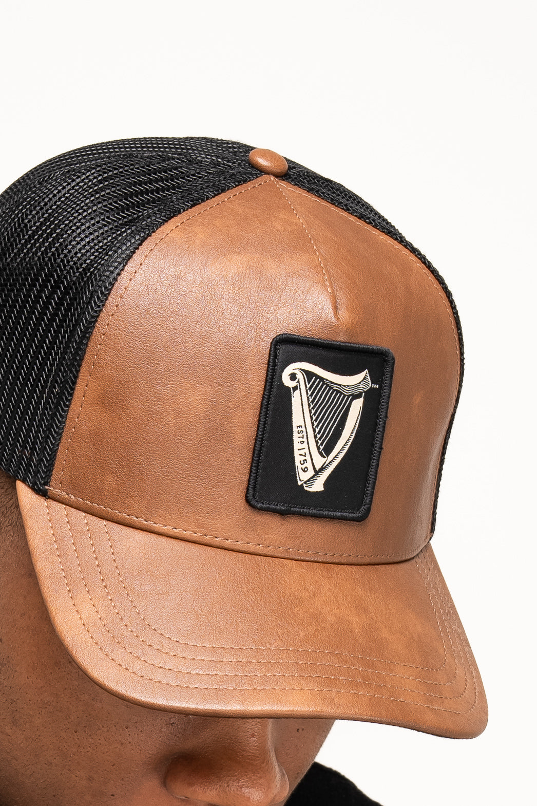 A person wears the Guinness Brown Leather Trucker Cap from Guinness Webstore US, featuring a mesh back, leather peak, and a front patch with the iconic Guinness harp logo.