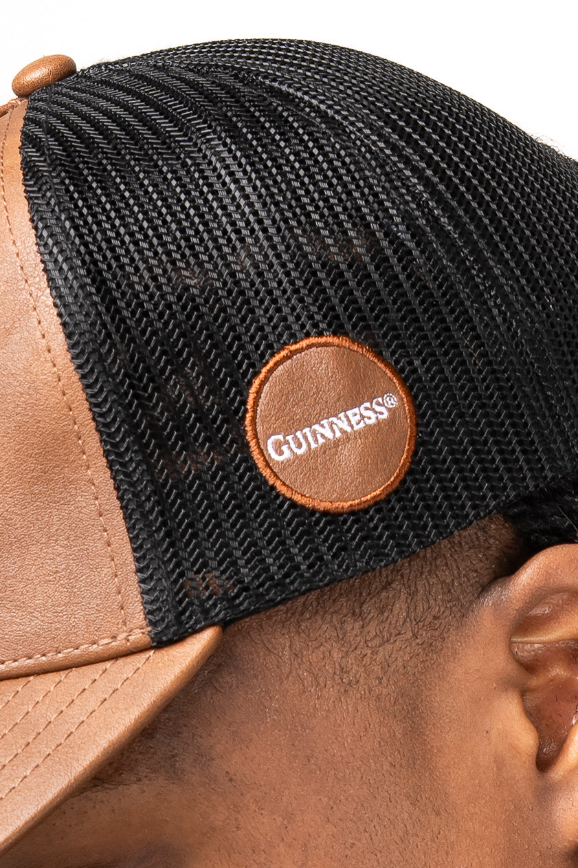 Close-up of a person wearing the Guinness Brown Leather Trucker Cap from Guinness Webstore US, featuring a leather peak and a round harp logo patch on the side.