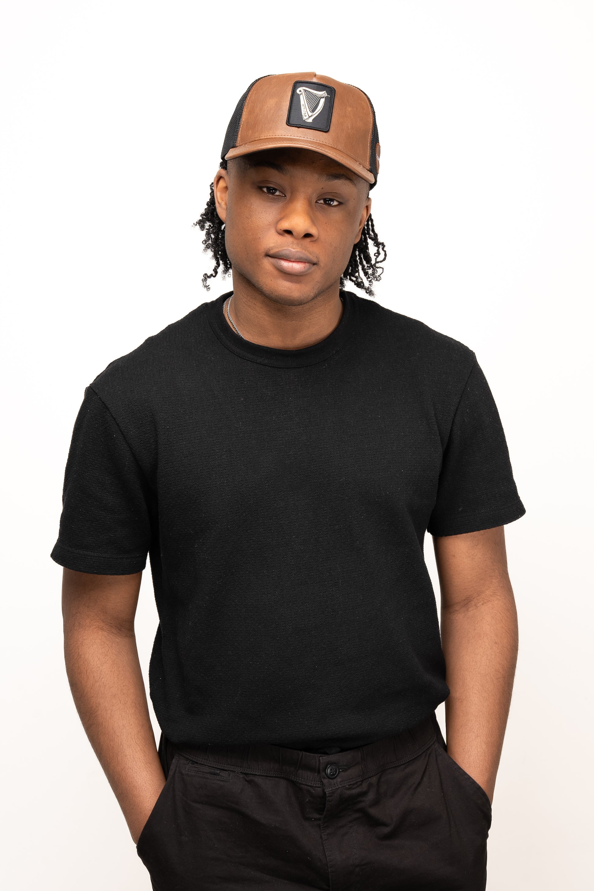 A person stands with hands in pockets against a plain white background, wearing a black t-shirt, black pants, and the Guinness Brown Leather Trucker Cap from Guinness Webstore US featuring a leather peak and snap-back closure.