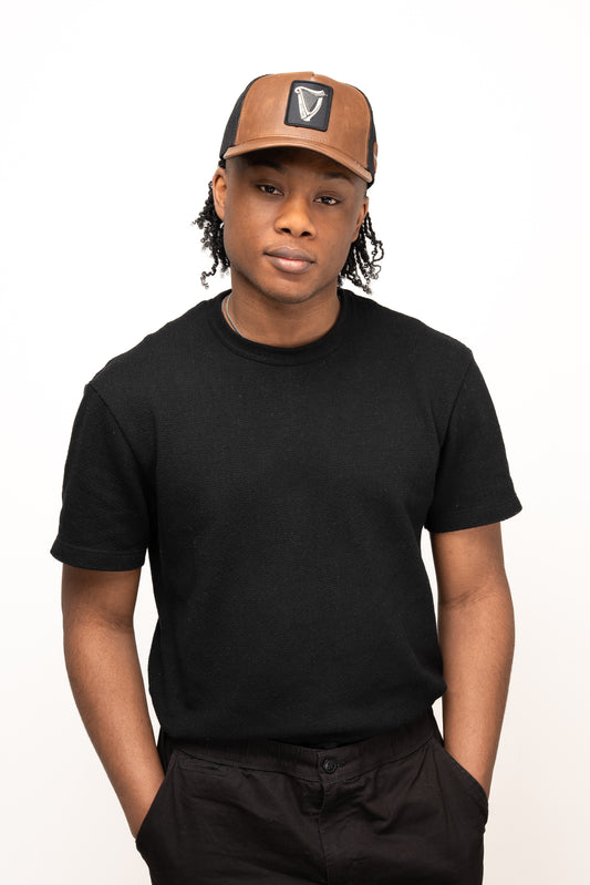 A person stands with hands in pockets against a plain white background, wearing a black t-shirt, black pants, and the Guinness Brown Leather Trucker Cap from Guinness Webstore US featuring a leather peak and snap-back closure.