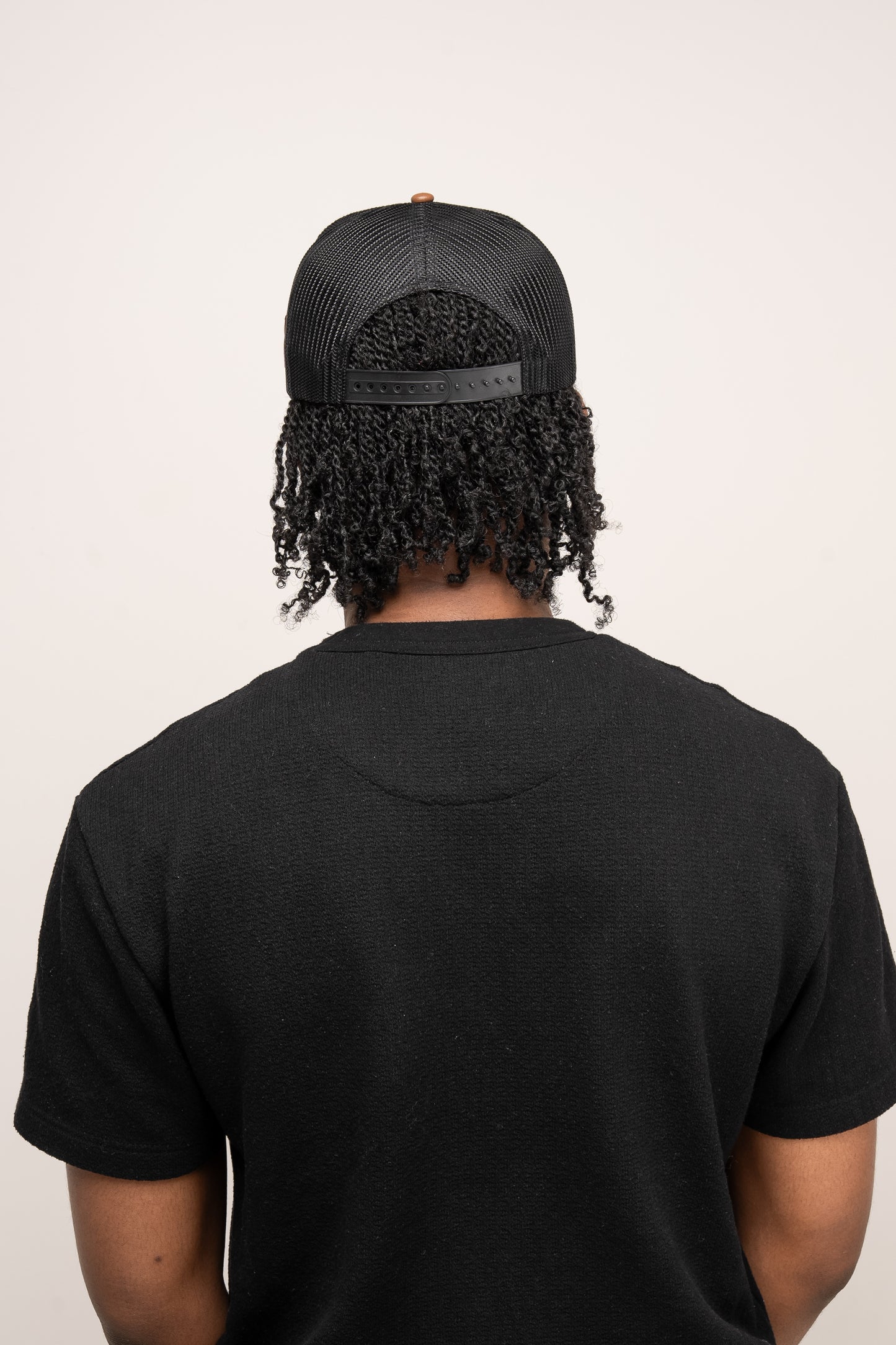 A person with curly hair in a black T-shirt wears the Guinness Webstore US Brown Leather Trucker Cap, featuring a leather peak, and stands facing away from the camera against a plain light background.