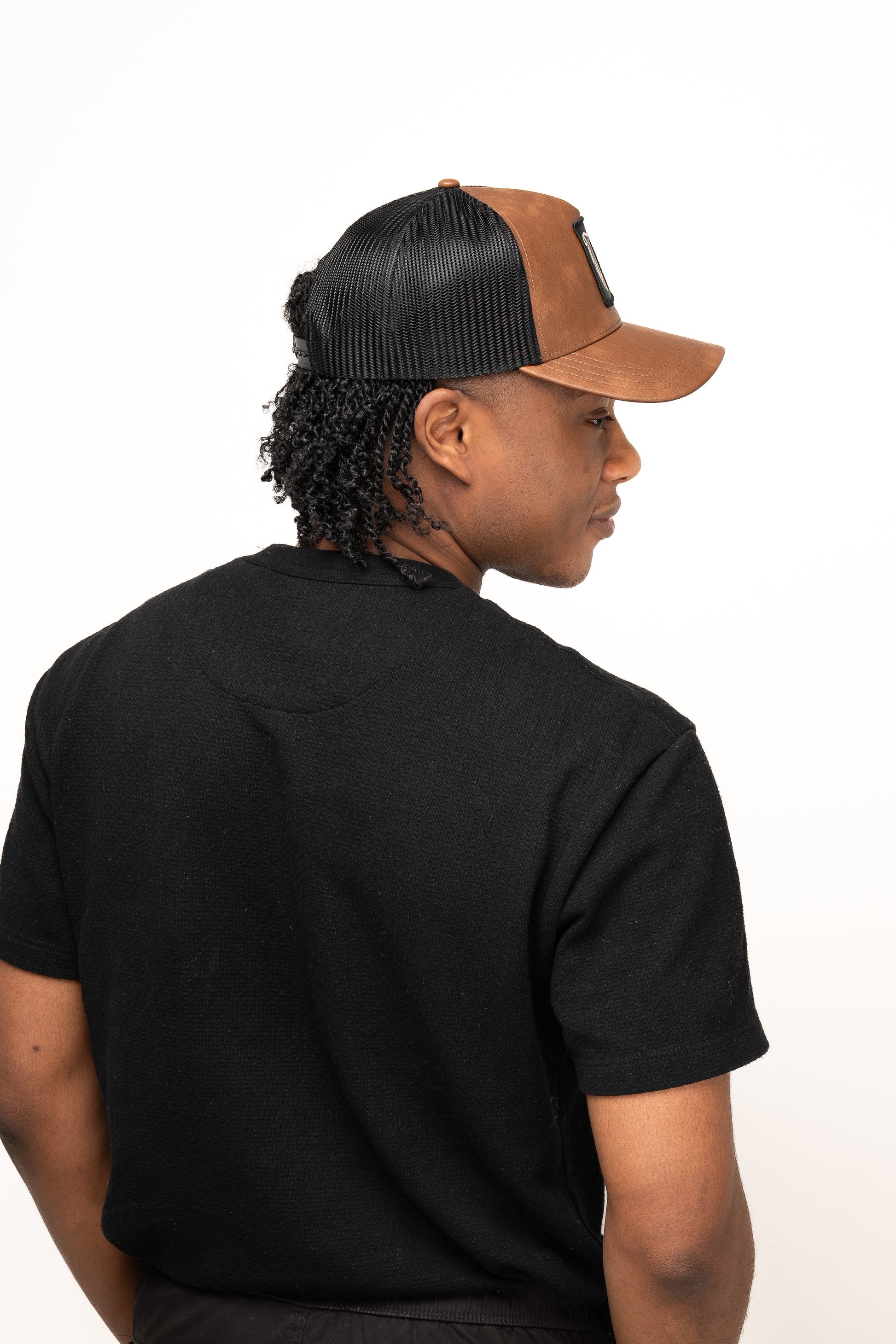 Someone wearing a black T-shirt and a Guinness Brown Leather Trucker Cap from Guinness Webstore US, featuring a leather peak and harp logo, stands with their back turned, looking sideways against a plain light background.
