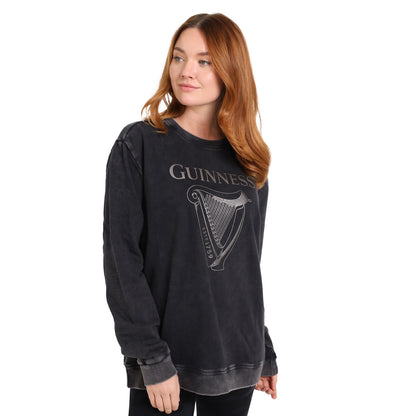 A woman with long red hair wears the Guinness Embossed Print Trademark Sweater from Guinness Webstore US, a loose black BCI cotton fleece sweatshirt with an embossed harp logo and vintage edge, standing against a plain white background.