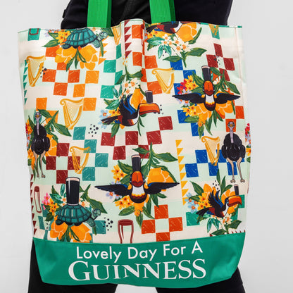 A person holds a Guinness Toucan Beach Bag from Guinness Webstore US, featuring toucans, harps, flowers, and “Lovely Day For A Guinness” text on a green Hawaiian-patterned background at the bottom.