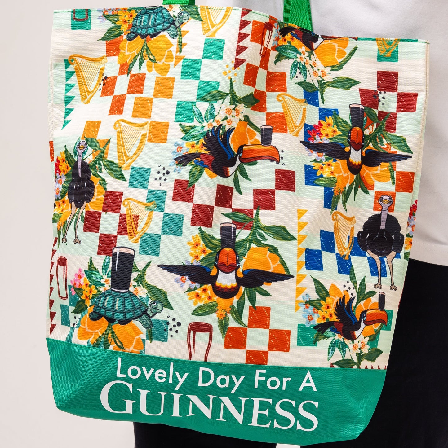 The Guinness Toucan Beach Bag from Guinness Webstore US features vibrant toucan and harp art, pint glasses, a Hawaiian pattern, and "Lovely Day For A GUINNESS" on a green border. Spacious 20L capacity.