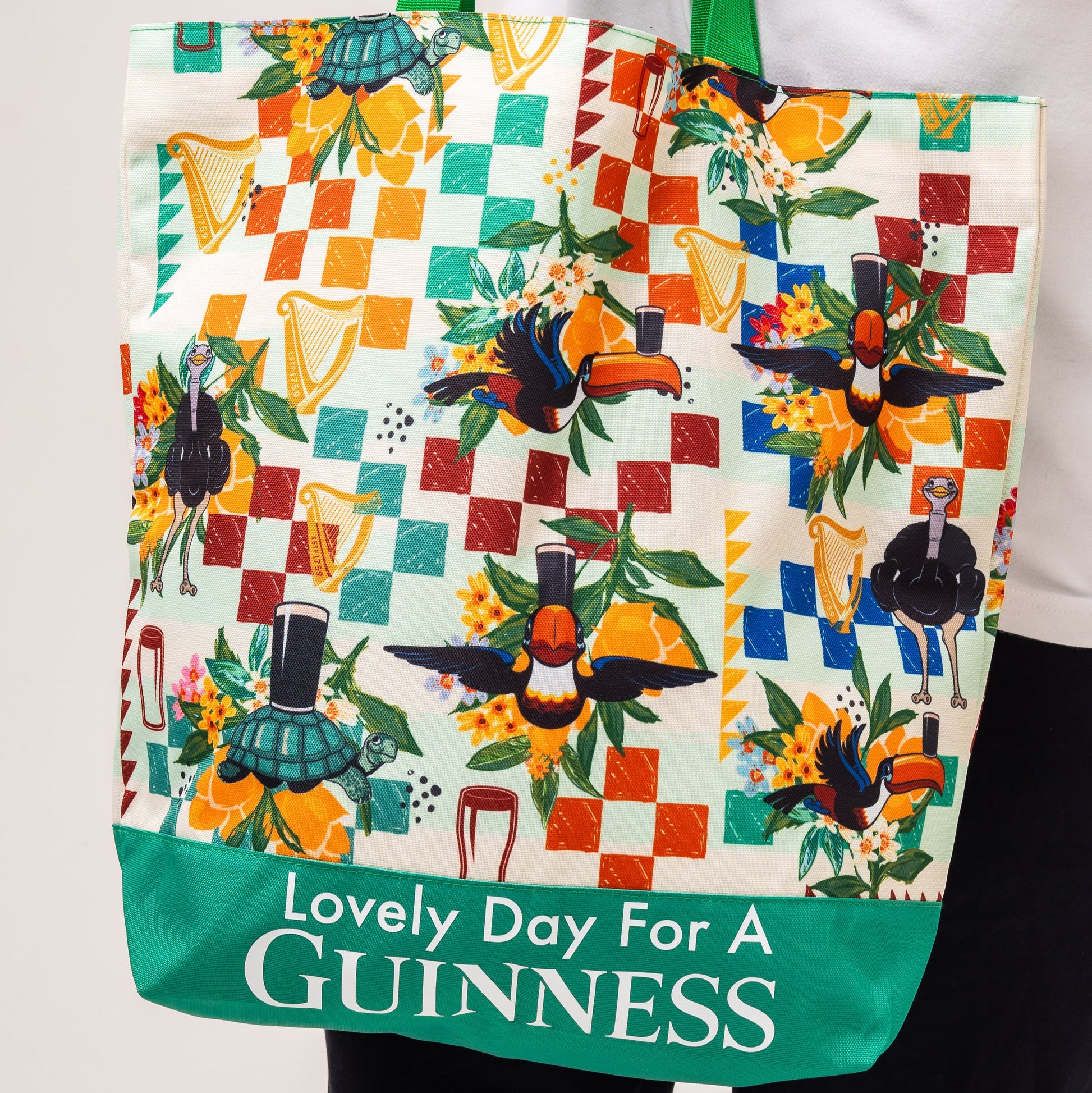 The Guinness Toucan Beach Bag from Guinness Webstore US features vibrant toucan and harp art, pint glasses, a Hawaiian pattern, and "Lovely Day For A GUINNESS" on a green border. Spacious 20L capacity.
