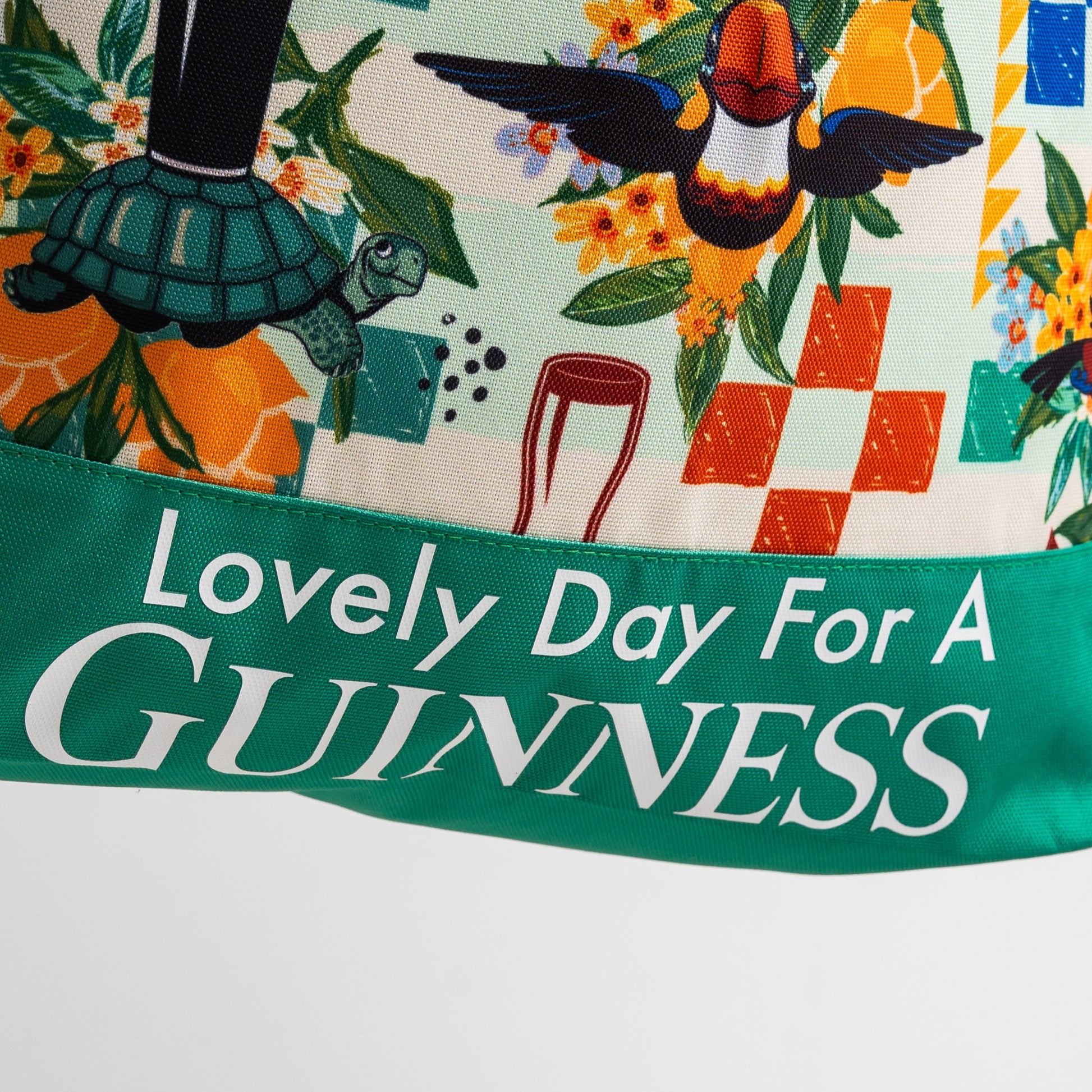 A vibrant illustration featuring a toucan, turtle, flowers, and "Lovely Day For A Guinness" text on green, inspired by Hawaiian patterns. Ideal for fans of the spacious 20L Guinness Toucan Beach Bag from Guinness Webstore US.