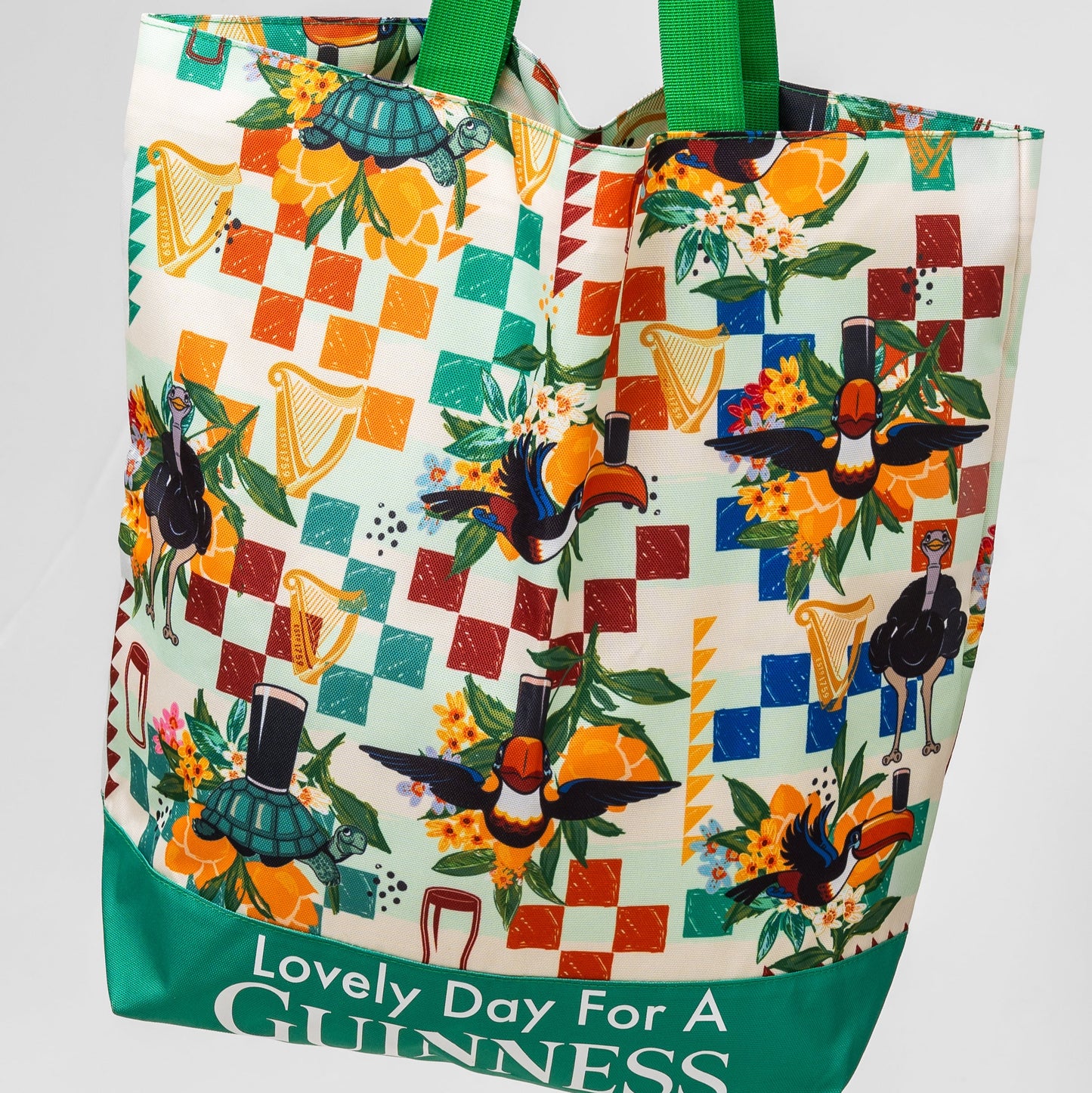 The Guinness Toucan Beach Bag from Guinness Webstore US features green handles, a 20L capacity, and vibrant branding with toucans, top hats, flowers, harps, and a colorful Hawaiian-themed background.