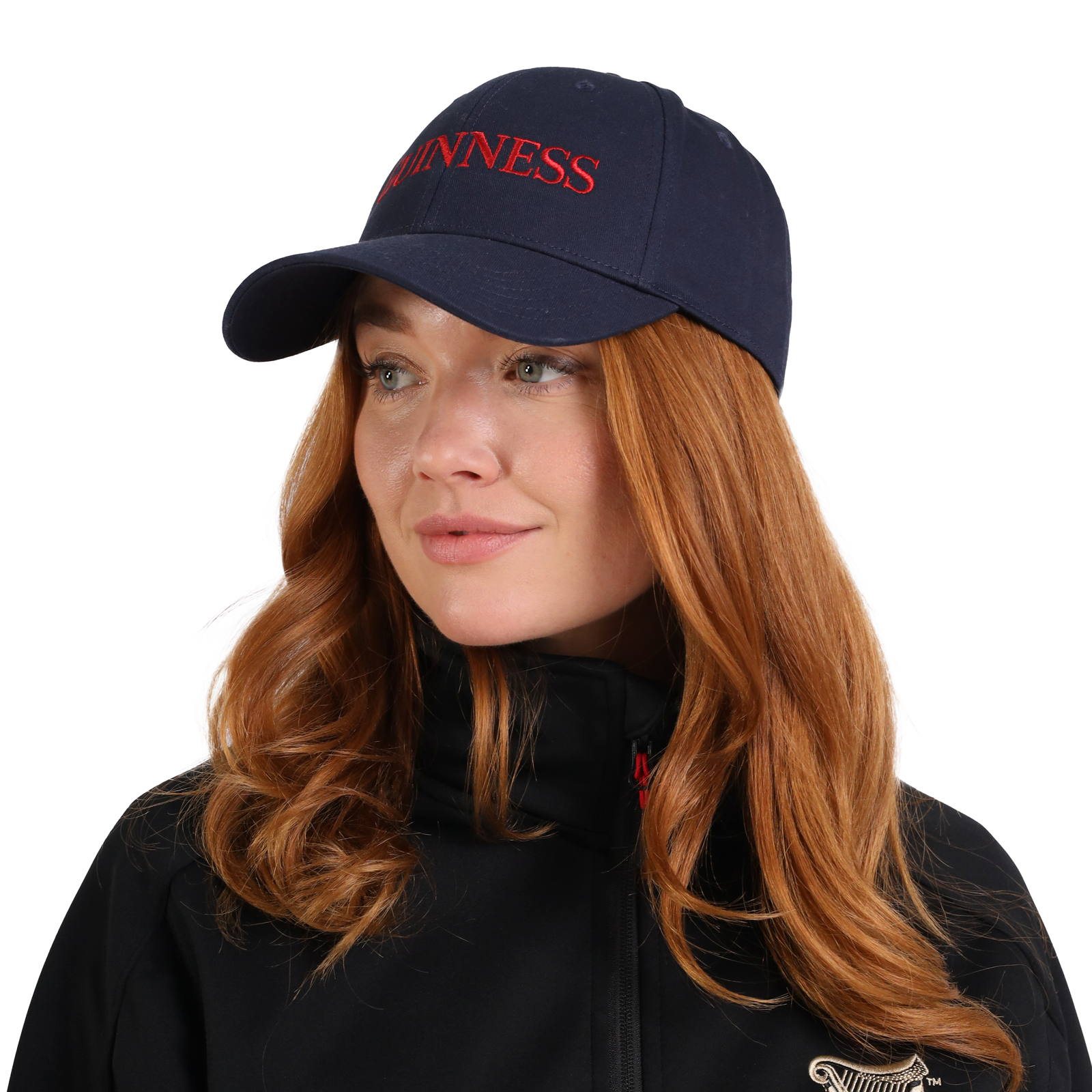 A person with long red hair wears a Guinness Baseball Cap from the Transport Collection, featuring navy blue with red lettering, along with a black jacket, looking slightly to the side.